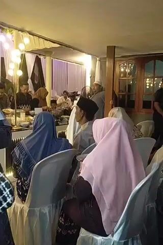 A group of people, primarily women wearing headscarves, are seated on white chairs at an indoor event. The room is decorated with white drapes and string lights. In the background, a man in a white hat and a grey shirt is speaking to someone. Other guests are visible, some at tables. The overall atmosphere appears to be a formal gathering or celebration.