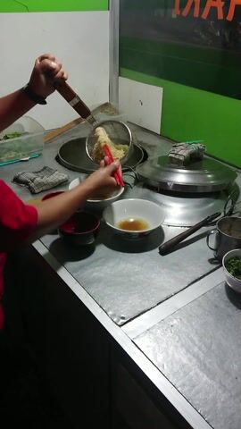 A person preparing food at a metal food stall. Using a metal strainer with a wooden handle, they transfer noodles from a steamer into bowls. Two red bowls and a small bowl of chopped green herbs are also on the stainless steel counter. The person wears a red shirt and a smartwatch. The stall is painted in green and white.