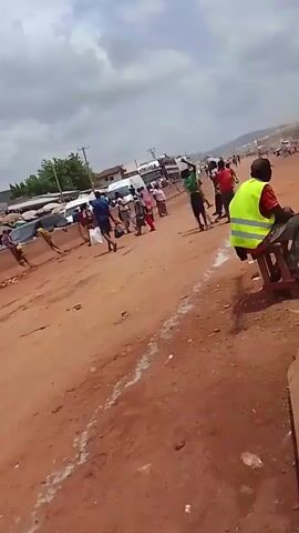 The video shows a crowded street scene in a developing country. The ground is red dirt, and there are several people walking and standing around. Many are wearing bright clothing, and some are carrying umbrellas. There are a few vehicles on the road, including a green van and some auto-rickshaws. Several people are wearing yellow safety vests. One man is sitting on a low stool, and another is working on something with his hands near some debris. In the background, there are several multi-story buildings. The sky is overcast.