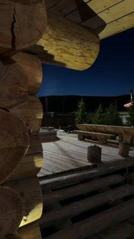 This video depicts an outdoor wooden deck at night, presumably part of a log cabin or a similar rustic structure in Drokino, Russia. In the foreground, large, hewn logs of a building frame the left side of the shot, illuminated by warm lights. The deck itself is made of wooden planks and features a round fire pit in the center, surrounded by wooden benches and tables. Small trees are visible beyond the deck, and a distant building with glowing string lights can be seen against the dark, clear night sky. The scene is quiet and still, with no people visible or specific activities taking place. In the background, male voices can be heard speaking Russian, with one clearly stating "Да, похуй вообще, блять" (Yeah, I don't give a shit, fuck) and mentioning "выпей пиво" (drink beer).