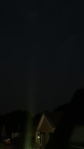 The video captures a nighttime scene with a dark sky. In the middle ground, silhouetted against the sky, are the roofs and upper portions of houses and trees. A faint, bright green light beam is visible originating from the bottom of the frame and extending upwards. At approximately 00:02, a flash of reddish-orange lightning illuminates the clouds in the distance, briefly lighting up the sky. The lightning flash is brief and subtle. The rest of the video shows the dark, uninterrupted sky.