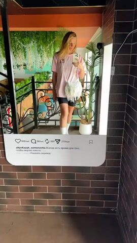 A woman with long blonde hair takes a mirror selfie in an indoor setting. She is wearing a light pink oversized t-shirt with a small graphic, black cycling shorts, white socks, and light-colored chunky sneakers. A white, fringed macrame-style crossbody bag with a braided strap is draped over her shoulder. She smiles gently at her reflection, holding a white smartphone (likely an iPhone) to capture the image.

The mirror she is posing in front of is designed to mimic an Instagram post. Below her reflection, an Instagram-like interface displays various elements: a like count of "956 тыс" (956k), comment and share icons, a save icon, and a view count of "270 тыс" (270k). The username `allynkazyk.semenovka` is visible, along with a caption in Russian that reads: "Всегда есть время для того, чтобы вкусно поесть." (Always there is time to eat deliciously.). Below the caption, it states "2 дня назад" (2 days ago) and "Показать перевод" (Show translation). A vertical LED light strip illuminates the right side of the mirror frame.

The background reflected in the mirror shows a lush indoor environment with hanging green plants, a black metal railing, and hints of an orange-colored wall or ceiling. A potted plant with large green leaves is visible to the right, and the wall surrounding the mirror is made of brown brick. The setting appears to be a cafe or a similar establishment, likely in Semenovka, Kyrgyzstan, as indicated by the Instagram username. The scene is well-lit, suggesting daytime.