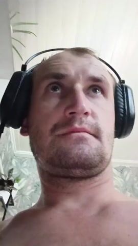 The video shows a close-up, low-angle shot of a man wearing black headphones. He is looking around, seemingly at something above and in front of him. His expression changes from a slightly surprised look to a more neutral one. The background appears to be a room with light-colored, paneled walls and a plant in the upper left corner. There is also some wallpaper visible behind him with a nature-themed pattern. In the lower left, a shelf is visible with some items on it. The man's shoulders are bare.