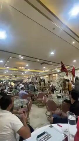 The video shows a large, brightly lit banquet hall with many round tables and chairs arranged throughout the space. People are seated at several tables, engaged in conversation or watching the central area. In the middle of the hall, a group of individuals is moving, some holding flags or ceremonial objects. The hall has a neutral color palette with gold accents, and there are decorative floral arrangements at some tables. The video is taken from a low angle, capturing the expansive interior of the hall.