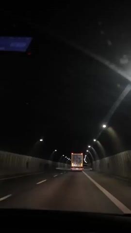 This is a first-person perspective video taken from inside a vehicle traveling through a dark tunnel. The tunnel has a curved ceiling and is illuminated by a series of overhead lights spaced at intervals. The road has clear white lane markings. Ahead, a yellow truck with its brake lights illuminated is visible in the distance, also traveling through the tunnel. The video captures the vehicle's movement deeper into the tunnel, with the truck gradually receding.