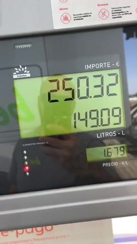 The image displays a close-up of a gas pump's digital screen at a fueling station in Torrent, Spain. The screen shows the current fueling transaction in progress.

**Subjects:** The primary subject is the gas pump display. Reflected prominently on the screen is a person, likely the photographer, wearing a dark t-shirt. The reflection clearly shows tattoos on the person's left arm and them holding a smartphone, indicating they are recording or photographing the display.

**Objects:** The gas pump display shows three main readings: "IMPORTE - €" (Amount - €) currently at 256.53, "LITROS - L" (Liters - L) at 152.79, and "PRECIO - €/L" (Price - €/L) fixed at 1.679. The brand "Petrotec" is visible in the top left corner of the display. Below the main readings are four indicator lights, three green and one red, next to the text "SUMINISTRO MÍNIMO: 2L" (Minimum supply: 2L).

**Scene/Setting:** The scene is an outdoor gas station during daylight hours, evidenced by the bright reflections on the screen.

**Activity:** The increasing numbers for "IMPORTE" and "LITROS" indicate that a vehicle is actively being fueled.

**Notable Details:** The language on the display is Spanish, confirming the location. Small informational signs above the main display provide rules: one for "Motos" (Motorcycles) stating "No puedes repostar con el motor encendido en el vehículo." (You cannot refuel with the engine running in the vehicle.), and another for "Envases" (Containers) stating "Solo puedes repostar en envases homologados y sin llenar más del 90% de capacidad." (You can only refuel in approved containers and without filling more than 90% of capacity.). The currency used is the Euro (€). Part of a "pre-pago" (pre-payment) sign is visible at the very bottom of the frame.