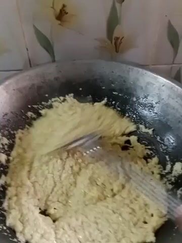 The video shows a close-up of a cooking pan where a light yellow, granular mixture is being stirred with a metal spatula. The mixture appears to be somewhat clumpy and is spread across the bottom of the pan. The background shows a tiled wall with a subtle floral pattern. The lighting is moderate, and the focus is on the food being cooked.