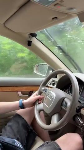 The video shows the interior of a car driving on a road in the rain. The camera is positioned behind the steering wheel, looking out the windshield. The steering wheel is visible, as well as the dashboard and the passenger side of the car. A fuzzy toy is hanging from the rearview mirror. A can of energy drink is in the cup holder. The windshield is wet with rain, and the wipers are not visible. Trees and a road are visible through the windshield. Other cars can be seen in the distance. The video is shaky, suggesting the car is in motion.