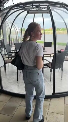 A woman with blonde hair pulled back in a ponytail walks into a clear, enclosed outdoor dining structure. The structure contains a round wooden table surrounded by six black chairs. The table is set with a menu and a napkin holder. The structure is located next to a lake, and buildings can be seen in the distance. The sky is overcast.