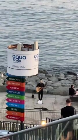 A street performer plays an acoustic guitar near a rocky shoreline. He is standing on a paved area facing the sea. In front of him is a signpost with multiple directional arrows indicating various businesses, such as "BOSS CAFE," "STARBUCKS," and "SUSHICO." Behind the performer, several people are seated on the rocks, some appearing to be watching him. To the right, two individuals are walking away, one of whom is carrying a backpack. In the foreground, a railing and a section of stairs are visible. The water behind the performer is calm, with gentle waves lapping against the rocks.