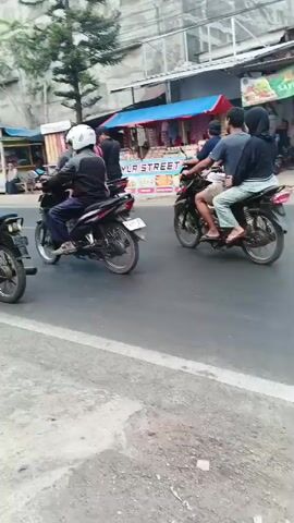 A street scene with several people riding motorcycles. A group of people in red and black uniforms are marching on the side of the road, carrying a large Indonesian flag. One person is riding a three-wheeled vehicle, possibly a motorcycle with a sidecar, which is also carrying people and the flag. The scene appears to be a procession or parade on a street lined with buildings and trees.
