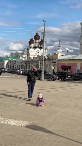 A woman and a young child are standing on a city square with historic buildings in the background. The woman is wearing a black leather jacket and blue jeans, and sunglasses. The child, wearing a pink and purple outfit with a hat, is initially on the ground but then stands up holding a small ball. A man on a green bicycle rides past, doing a wheelie. There are cars parked along the side of the square, and the sky is partly cloudy.