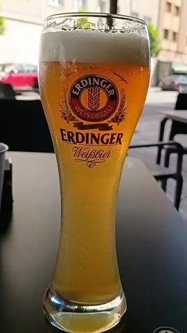 A close-up shot focuses on a tall glass of Erdinger Weissbier, filled to the brim with frothy white foam and golden beer. Condensation beads on the outside of the glass, indicating it is chilled. The beer is set on a dark table, with the background slightly blurred. In the out-of-focus background, a pink car is visible on a street, and the side of a building with windows can be seen. Two black chairs are also present in the background, suggesting an outdoor seating area, possibly a cafe or restaurant. The lighting appears to be natural daylight. There are no people visible in the frame.