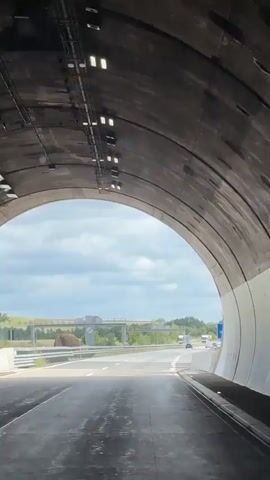 The video begins inside a tunnel and then moves outside into daylight, showing a highway with multiple lanes of traffic. The sky is cloudy. Several trucks and cars are visible on the highway. A large brown object that appears to be a stuffed animal or mascot is positioned on the side of the road near the left lane. There is also a blue sign on the right side of the road with a white 'P' indicating parking. The tunnel interior is concrete with overhead lighting fixtures.