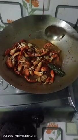 A wok filled with crab claws is being stirred by a ladle and a spatula on a stovetop. The crab claws are reddish-orange and appear to be cooked. The wok is made of metal and has a dark interior, suggesting it has been used for cooking. The stovetop is also metallic and has a burner visible beneath the wok. In the background, there is a wall with a tiled pattern. The lighting is dim, and the overall scene suggests a home kitchen environment where food is being prepared.