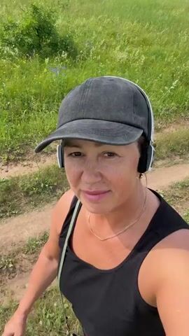 A woman wearing a black baseball cap and black tank top walks on a dirt path through a grassy field. She has headphones on and a gold necklace. The path curves to the right and continues uphill. Trees and some buildings are visible in the distance. The sky is cloudy.