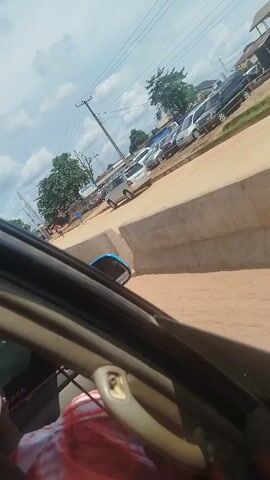The video shows a street scene in Okha, Nigeria. The camera is positioned inside a car, looking out the passenger side window. Outside, there are several cars parked along a dirt road, and a concrete barrier runs alongside the road. A person on a motorcycle is visible riding down the road. In the background, there are buildings and trees, with power lines overhead. The sky is cloudy, suggesting it is daytime. The overall impression is of a typical street scene in a town or city. There is no discernible emotional content, and the activity appears to be normal daily life.

One visible text detail is the word "SENNA" printed on a car window in the foreground. There is also partial text visible on the car door handle which is difficult to read.