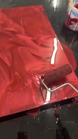A hand holding a small paint roller with a red handle is rolling a swirling red liquid on a flat, dark surface. The liquid has a metallic sheen and is creating patterns of lighter and darker red. There are some drips of the red liquid on the dark surface. In the upper right corner, there is a small white container with a red lid and a wooden stick. The container has text on it, including "measures". The action appears to be part of an art or craft project.