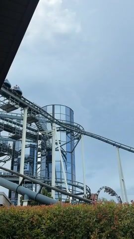 A roller coaster is shown moving from left to right along a track. Several cars are visible on the track, each carrying passengers. In the background, a tall, cylindrical building with reflective blue glass windows stands beside the roller coaster. A dense green hedge is in the foreground, obscuring the lower portion of the scene. The sky is partly cloudy.