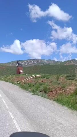 A video shows a monument on a hill. The monument is a sculpture of a horse on a pedestal. The monument is reddish-brown. The hill is covered in green grass and some red dirt. There is a blue sky with white clouds. A road is in the foreground. The video pans to the right, showing more of the hill and mountains in the background. Some animals, possibly horses, are seen grazing on the hillside.