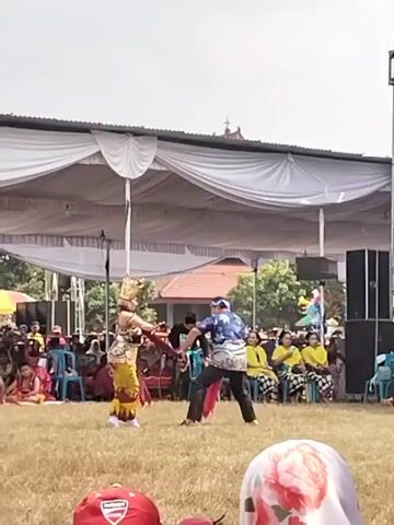 A staged outdoor performance is taking place under a large white tent. Two performers are on a grassy field. The performer on the left wears traditional yellow and red attire with a headdress. The performer on the right wears a blue and white patterned shirt, dark pants, and a headscarf. They appear to be engaged in a choreographed dance or fight, with a red sash held between them. A person in dark clothing stands behind them. An audience is seated on chairs behind the performers, some wearing yellow and patterned clothing. The background shows trees and buildings. In the foreground, parts of audience members' heads and hats are visible, with one red hat in the lower left and another head with a floral patterned scarf on the right. The sky is bright and overcast.