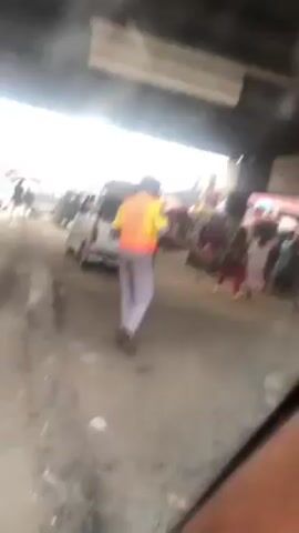 The video shows a street scene under a bridge, with a man in a yellow and orange vest standing in the foreground. He is wearing light grey trousers. In the background, there are cars, motorcycles, and people moving around. A man in black rushes across the street to the left of the central figure. The environment appears to be an urban area with signs of congestion and activity. The video is shot from inside a vehicle, looking out.