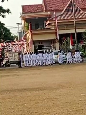 A group of people in white uniforms are walking in a line on a dirt road in front of a building decorated with red and white flags. The people are walking from left to right. There are cars and motorcycles on the side of the road. Trees and other foliage are visible in the background. The weather appears to be overcast.