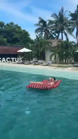 A woman is floating on a red-and-white striped inflatable raft in clear turquoise water. She is lying on her back, wearing a swimsuit and sunglasses. She raises her right arm, then her left leg into the air. The background includes a sandy beach with lounge chairs, palm trees, and a building with the word "CACTUS" in large letters. The sky is blue with some clouds.