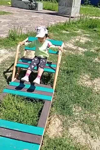 A child wearing a light green long-sleeved shirt, black leggings with a white pattern, white socks, white shoes, and a white baseball cap is sitting on a wooden structure. The structure appears to be a slide or ramp with teal and brown planks. The child is holding onto the sides of the structure and looking forward. The child is on a grassy area with some bushes and a paved path in the background. There is also a stone monument in the background. The scene is lit by sunlight.