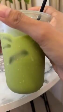 A hand is holding a clear plastic cup filled with a green beverage and ice cubes. The cup has condensation on the outside. A black straw is visible in the cup. The cup is sitting on a white table with a marble-like pattern. In the background, a glass vase with greenery is visible. The hand is turning the cup.