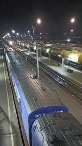This video shows a nighttime view of a train yard. The foreground features the roof of a blue train, with "POCHTA ROSSII" (Russian Post) written on it. In the middle ground, several tracks are visible, occupied by numerous yellow tank cars. The tank cars are illuminated by overhead lights, creating bright spots and casting shadows. In the background, more train cars and structures are dimly lit. The scene appears to be a stationary train yard at night, with the focus on the postal train in the foreground and the parked tank cars beyond.