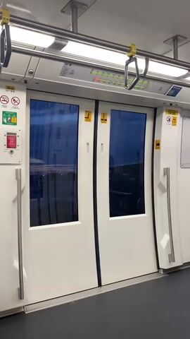 The camera is held stationary, providing an interior view of a train car. The focus is on a pair of closed, white-framed doors, with dark-tinted windows that offer a glimpse of the outside environment. The sky visible through the windows suggests either dawn or dusk. Overhead, metal bars run the length of the train car, equipped with yellow handholds for passengers. Various informational stickers and a red emergency button are affixed to the wall adjacent to the doors. The lighting inside the train car is bright and even. There are no people visible in the video. The scene is static and devoid of action.