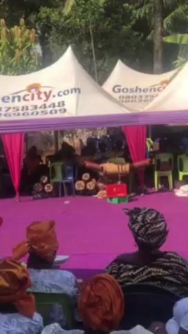 Here's a description of the video:

The video shows a gathering of people under several white tents with pink accents. The tents are labeled "Goshencity.com" along with a phone number. A stage is set up at the front, covered in purple fabric. A band of musicians is visible on the stage, playing traditional instruments. Some of the instruments are displayed on the stage, while others are being played by the band. 

The audience sits on chairs, facing the stage. Most attendees wear traditional African attire, with many women sporting headwraps, including a group wearing orange headwraps. Some food and other items are arranged on small tables between rows of seating. 

The setting appears to be outdoors, with trees visible in the background. The atmosphere is festive and celebratory.
