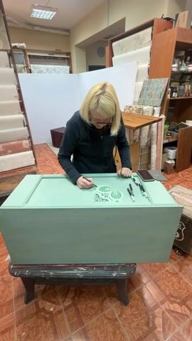 A blonde woman with glasses is painting a light green chest with markers. She is focused on the task, and her hands are holding a marker as she draws a design on the lid of the chest. There are several markers, a phone, and a tablet on the chest. The chest is on a wooden stand. The background of the scene is a room that appears to be a workshop or store, with shelves stocked with items, a carpeted staircase, and other furniture. The floor is tiled with a terracotta color.