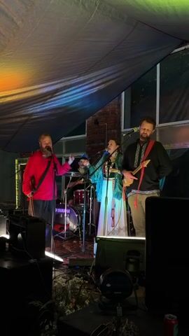 A live music performance is underway. Three musicians are visible on a stage under a dark, draped ceiling illuminated by colorful lights. A man in a red shirt and dark pants stands on the left, singing into a microphone and holding what appears to be a guitar. In the center, a woman in a blue and white ornate outfit, also wearing a microphone headset, sings expressively. To the right, a man in a black shirt with intricate embroidery and dark pants plays a stringed instrument. Behind them, a drummer is partially visible. The stage is set up with musical equipment, including speakers and monitors. The environment suggests an indoor venue with brick walls and a window visible in the background. The lighting is dramatic, with a spectrum of colors cast across the scene.