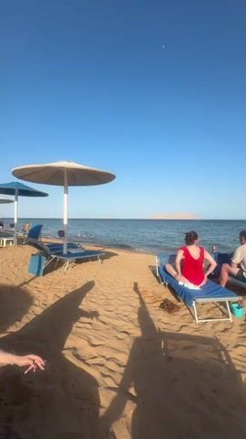 The video shows a sunny beach on a clear day. In the foreground, a person's shadow points towards a couple relaxing on blue beach chairs. A white umbrella is nearby. Further along the beach, a woman in a black wetsuit stands near a child who is lying down in the sand close to the water's edge. A long pier extends into the water. Some people are swimming in the sea. In the distance, a land mass can be seen on the horizon.