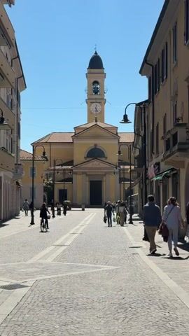 A pedestrian street, paved with brick and stone, leads toward a yellow church with a clock tower in the distance. Several people are walking down the street. There are shops on either side of the street. A cyclist rides toward the church. The sky is bright blue.