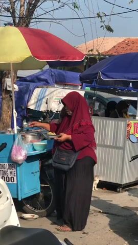 A woman wearing a maroon hijab and a maroon shirt is standing in front of a food cart. She is holding a white plate with food and a plastic bag. The food cart has a blue cart with a yellow and red umbrella. The cart has text on it that is partially visible. Behind her, there is another food cart with a blue umbrella and some people are visible inside. A cat is visible near the food cart behind her. The environment appears to be an outdoor market or street.