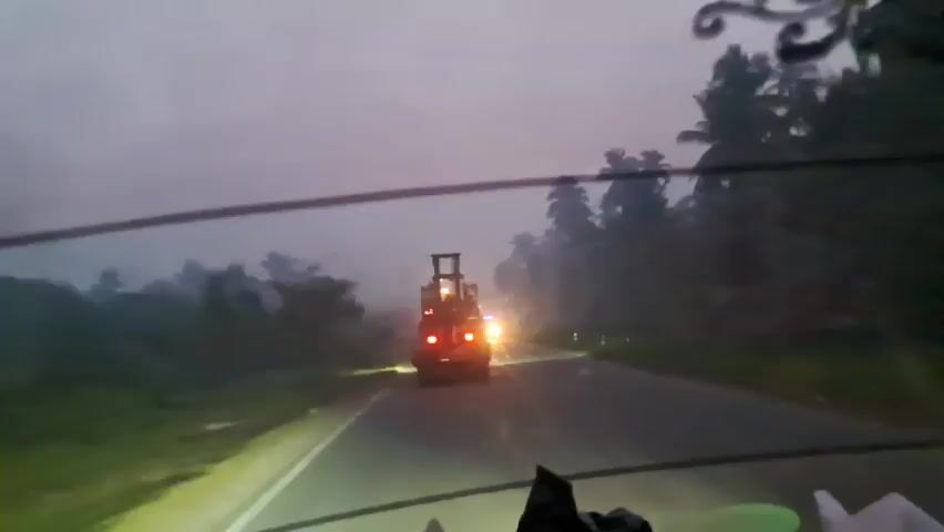 The video shows a view from inside a vehicle, looking out through the windshield. The vehicle is on a road in what appears to be a tropical environment, with palm trees visible on either side. The sky is overcast and the light suggests it is either early morning or late evening, possibly during rain or fog, as the visibility is low and there is a haze. In front of the vehicle, further down the road, are two vehicles. The lead vehicle appears to be a truck or large vehicle with its brake lights illuminated. Above the brake lights, there is a raised structure, possibly part of the cargo or equipment being transported. In front of this vehicle, slightly to the right, is another vehicle with bright headlights, possibly an accompanying vehicle or another vehicle on the road. The road is paved and has a white line marking the edge and a yellow line dividing lanes. The headlights of the vehicle the camera is in are also illuminated and are casting light onto the road and the surrounding vegetation. The overall atmosphere is dim and slightly eerie due to the low light conditions and haze.