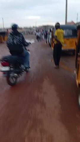 The video shows a muddy road in what appears to be a developing country. Several vehicles, including motorcycles and auto rickshaws (tuk-tuks) are present. A man wearing a yellow shirt and dark pants walks along the road. Puddles of muddy water are visible, indicating recent rain or poor drainage. The sky is overcast. There are several other people present, some standing and others riding motorcycles or in tuk-tuks.