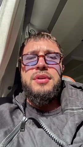 A man with a beard and glasses is looking at the camera. He is wearing a black jacket and a black shirt. The background appears to be the interior of a vehicle, with a window and a curtain. The lighting suggests it is daytime.