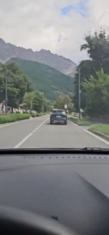 The video begins from the perspective of a driver in a car, showing a road ahead. A dark blue SUV is traveling in the same direction, in front of the car. The road is lined with trees and buildings on the left, and trees and a street light on the right. In the background, a large mountain range with green slopes and rocky peaks is visible under a cloudy sky. The car continues to drive down the road, passing the buildings and trees. The mountain scenery remains in the background. The camera is focused on the road ahead and the SUV.