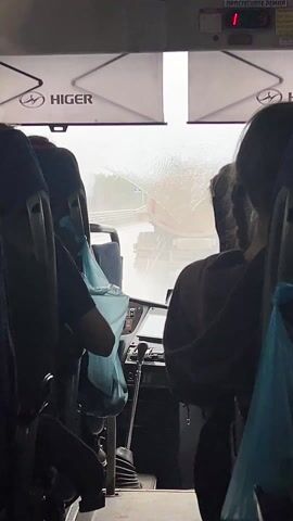 The view from inside a moving Higer bus looks toward the windshield, where wipers are in use due to heavy rain obscuring the view.  Other passengers are visible, their backs to the camera, and several plastic bags hang from the seats in front. The driver's area is partially visible, and a digital display above the windshield alternates between the numbers 4 and 09.  Outside, a red truck is visible through the rain.