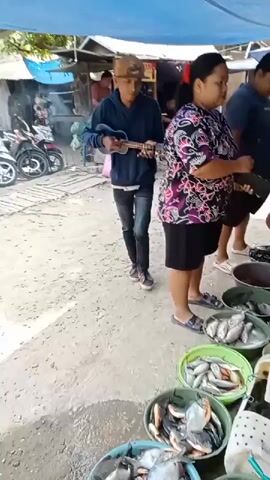 A young male wearing a blue hoodie, black jeans, a baseball cap, and sneakers is playing a ukulele. He is standing in a market, likely outdoors, with a blue tarp overhead. To his right, there are stalls with various goods, including baskets of fish for sale. Several people are present, including a woman in a black and purple patterned shirt, a man in a dark blue shirt, and another man in a blue cap and green shorts who appears to be interacting with the fish seller. Motorbikes are parked in the background. The ground is dirt and appears wet in places. The overall atmosphere is that of a bustling local market.