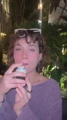 A young woman with curly brown hair and sunglasses on her head is outdoors at night, wearing a purple sweater and earrings. She is holding a small container with a white lid, taking a bite or sip from it, then looking up at the camera. The background includes plants and dim lighting.