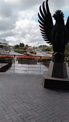 A black, statue of a bird with outstretched wings stands on a pedestal. The statue is positioned in the foreground, with a cobblestone pathway in front of it. Beyond the statue, a river flows, with planters filled with red flowers lining its banks. In the background, there is a bridge, buildings, trees, and a cloudy sky. The video pans slightly to the right.