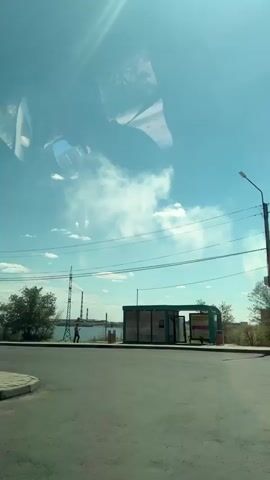 The video is a first-person perspective from inside a vehicle, looking out the windshield on a sunny day. The sky is a clear blue with some wispy white clouds. Reflections of the vehicle's interior, including what appears to be part of the dashboard and headliner, are visible on the windshield.

In the middle ground, there is a paved road and sidewalk. To the right, there is a bus stop shelter with a green roof and transparent walls. Behind the bus stop, there is a yellow advertising sign. Further in the distance, there is a body of water, and beyond that, a low-lying urban or industrial landscape with several tall structures, including what appear to be smokestacks or industrial towers, and some trees. A person is walking along the road in the distance.

A yellow and white bus is visible approaching and then passing by the trees on the left side of the frame. There are power lines running horizontally across the scene. The overall environment appears to be a somewhat undeveloped or industrial area with a bus stop.