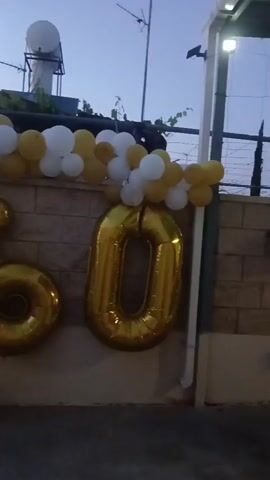 The video shows a celebration display with large gold balloons forming the number 60. These are situated below a garland of white and gold balloons that is draped over a wall. The wall appears to be made of textured stone or brick. In the background, there is a rooftop with a water tank and an antenna. The sky is visible, suggesting it is either dawn or dusk. The overall setting is outdoors, possibly a patio or yard. There are no people or explicit actions depicted in the video.