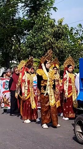 A group of people are walking in a procession on a paved road. They are dressed in elaborate traditional clothing, including ornate headdresses, embellished robes, and flowing sashes. The people are carrying flags and banners, some of which feature images and text. In the background, there are trees and foliage, suggesting an outdoor, possibly rural, setting. The sky is visible, indicating it is daytime. A blue vehicle is partially visible on the right side of the frame, and part of a tire is visible in the bottom right corner. Some of the people are waving.