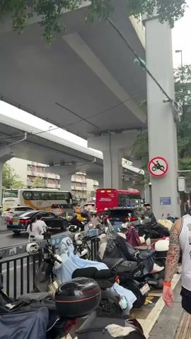 The video captures a street scene beneath an elevated highway, likely in an urban environment. A considerable number of parked motorcycles and scooters occupy the foreground, many covered with tarps or fabric. Some of these vehicles appear to be electric scooters. A street barrier separates the parked vehicles from a busy road where cars and buses are navigating through traffic. A large white bus with "VIP" branding is visible, along with a red bus and several cars. People are seen on some of the motorcycles, either riding or preparing to ride. One person in the foreground is interacting with their phone. A traffic sign prohibiting motorcycles is prominently displayed on a concrete pillar supporting the elevated highway. The overall atmosphere suggests a typical day in a densely populated city with significant vehicular traffic and reliance on two-wheeled transportation. The weather appears overcast.